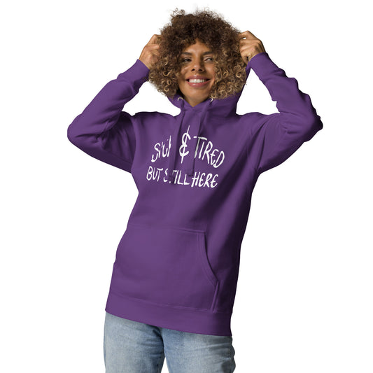 Person wearing a purple hoodie with text SICK AND TIRED BUT STILL HERE on a white background