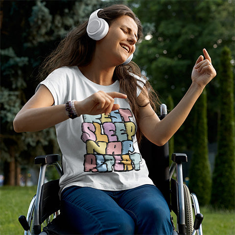 Person in a wheelchair wearing headphones and a colourful t-shirt with text REST SLEEP NAP REPEAT, sitting outdoors.