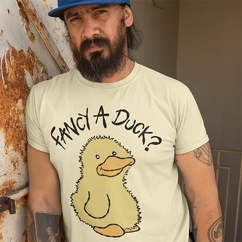 Man wearing a t-shirt with a duck graphic and text FANCY A DUCK?, standing against a rustic wall.